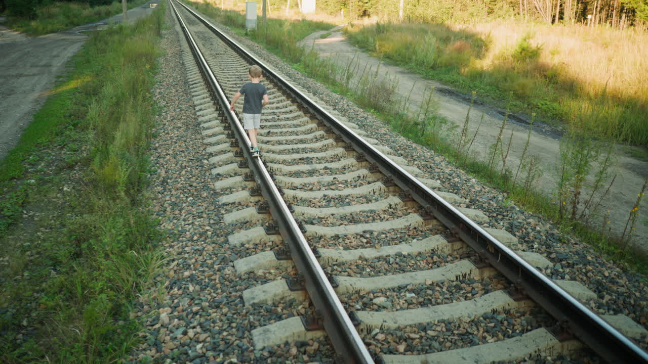 back view of little boy walking on rail beam in rural setting with gravel roads, grass, poles and forest edge under golden sunlight, showcasing balance, curiosity and exploration on warm summer day