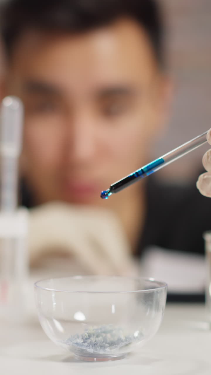 Young employee with latex gloves drops dark blue liquid into bowl with crystals doing experiments in scientific laboratory close view