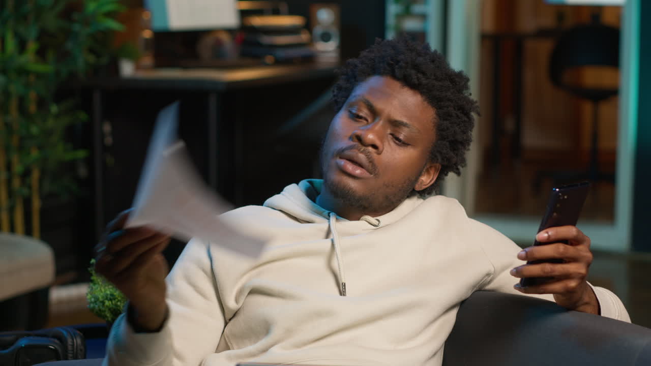 Black freelancer does his research by reading articles and data driven papers at home, using phone and pc to collect information for a new project. Man multitasking and studying files. Camera A.