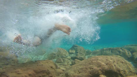 joven nadando en azul claro bajo el agua de la cascada con muchas rocas desde un ángulo plano