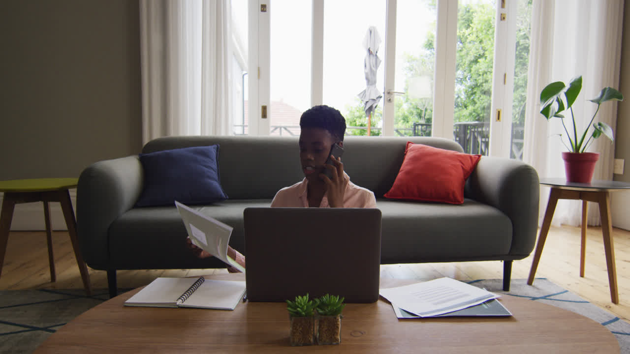 African american woman holding a document talking on smartphone while working from home