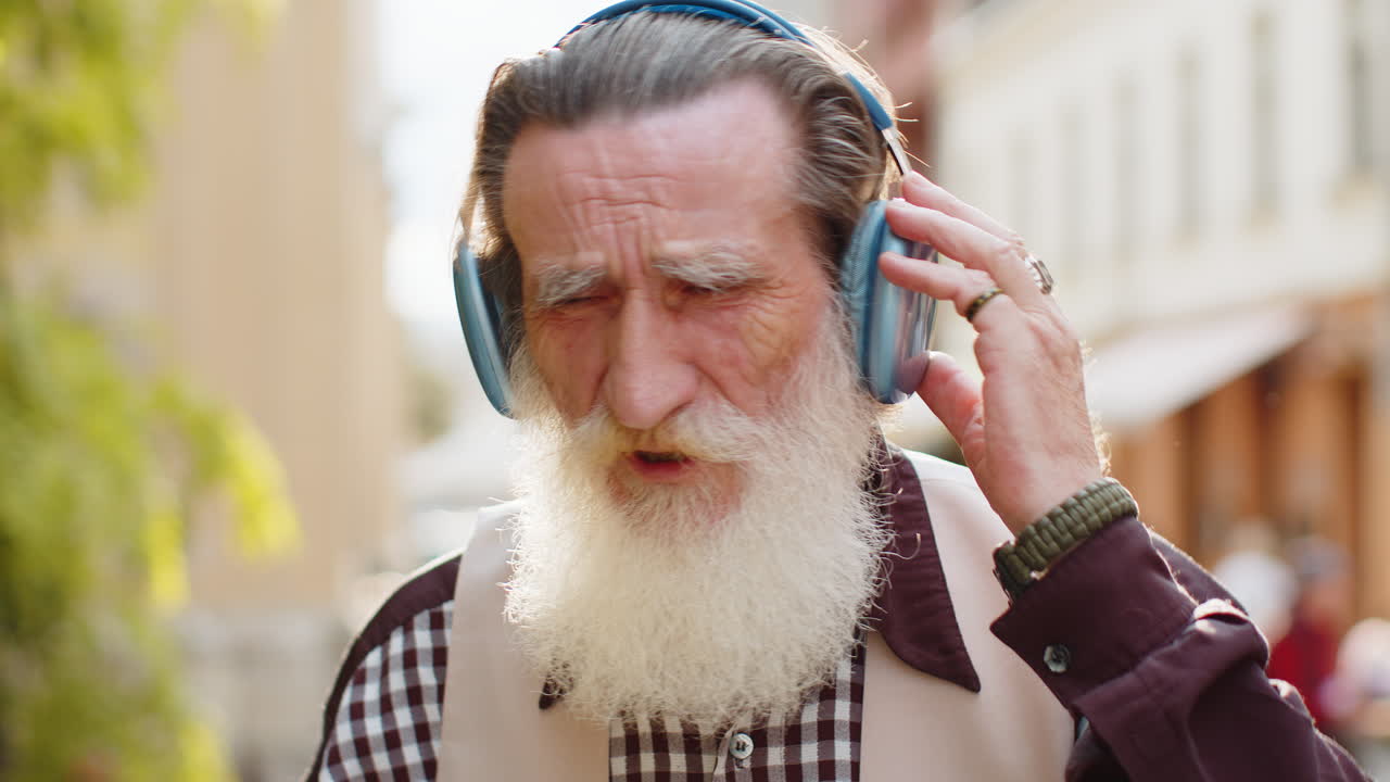 Happy senior old man in wireless headphones choosing listening music dancing outdoors city street