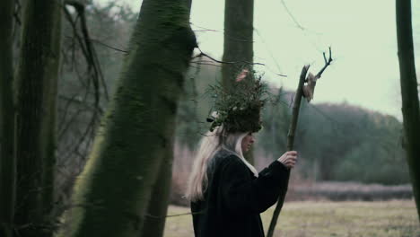 Wild-girl-with-natural-crown-and-skull-on-stick-walking-in-forest,-side-view