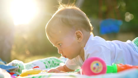 half a year old baby lies on the ground in sun beams in summer. kid interest in a toy moves it closer. side view.