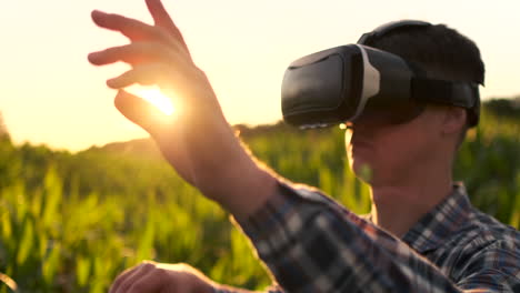 A-male-farmer-in-virtual-reality-glasses-wearing-a-plaid-shirt-and-jeans-in-a-field-at-sunset-controls-the-corn-irrigation-system.-Modern-farmer-new-technology-farmer-of-the-future