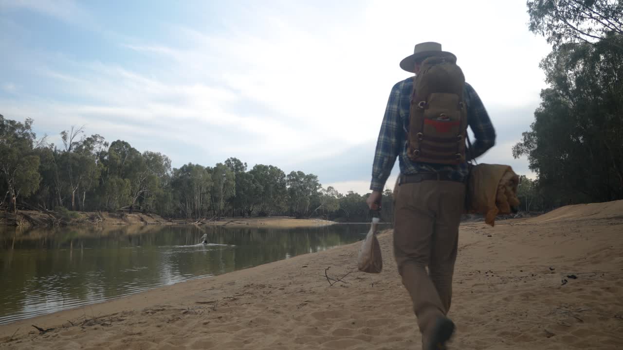 Traditional swagman walking along the banks of the Murray river with a pelican floating past.