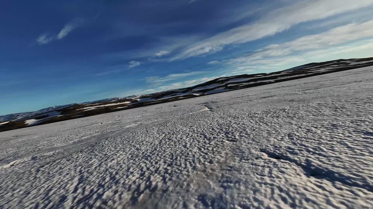 Un drone FPV sfiora a bassa quota la superficie ghiacciata del lago Heiðarvatn vicino a Egilsstaðir, sulla strada per Seyðisfjörður, sfrecciando su crepe ghiacciate sotto cieli di un blu intenso e montagne invernali.