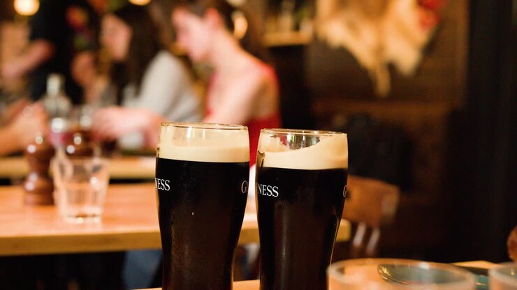 Una mano si allunga per prendere una birra scura stout su un tavolo di legno in un pub affollato e illuminato da una luce calda
