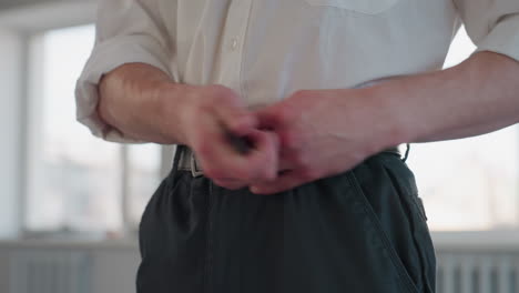 Portrait view of man hands fastening black belt around waist against bright window background conveying attention to detail and focused morning dressing routine with rolled sleeves in natural light