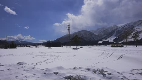 Amidst-the-captivating-winter-scenery-of-Hakuba,-Japan,-a-shot-captures-the-presence-of-electrical-pylons-towering-against-the-cloudy-sky,-while-a-car-gracefully-cross-the-snowy-field-in-the-frame