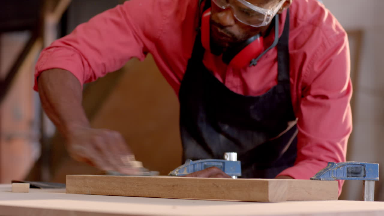African American man wearing goggles using sanding block on plank at workbench, with blue clamps