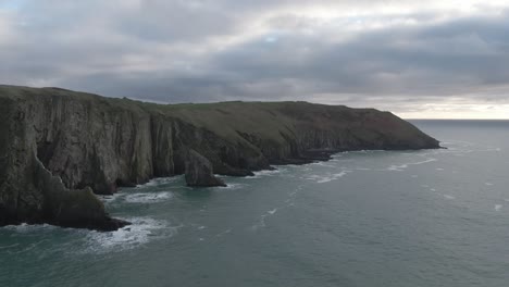 stationary aerial view of sea side cliffs with rough seas and waves hitting the cliffs, steep cliffs with cloud cover