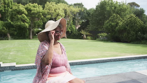 feliz anciana afroamericana en sombrero de sol sentada en el sol por la piscina, espacio de copia, cámara lenta