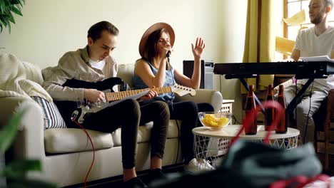 handsome young guitarist is playing looking at music sheet while vocalist is singing in microphone and keyboarder is playing the keyboard. musical band is rehearsing.