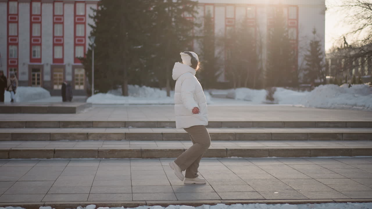 Youth in winter coat and headphones spins gracefully on snowy step pavement during golden hour, immersed in dance while people walk in background near trees and historic urban buildings