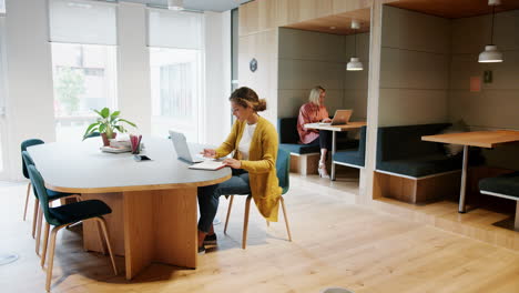 dos mujeres millennial trabajando en computadoras portátiles sentadas en una oficina creativa, longitud completa
