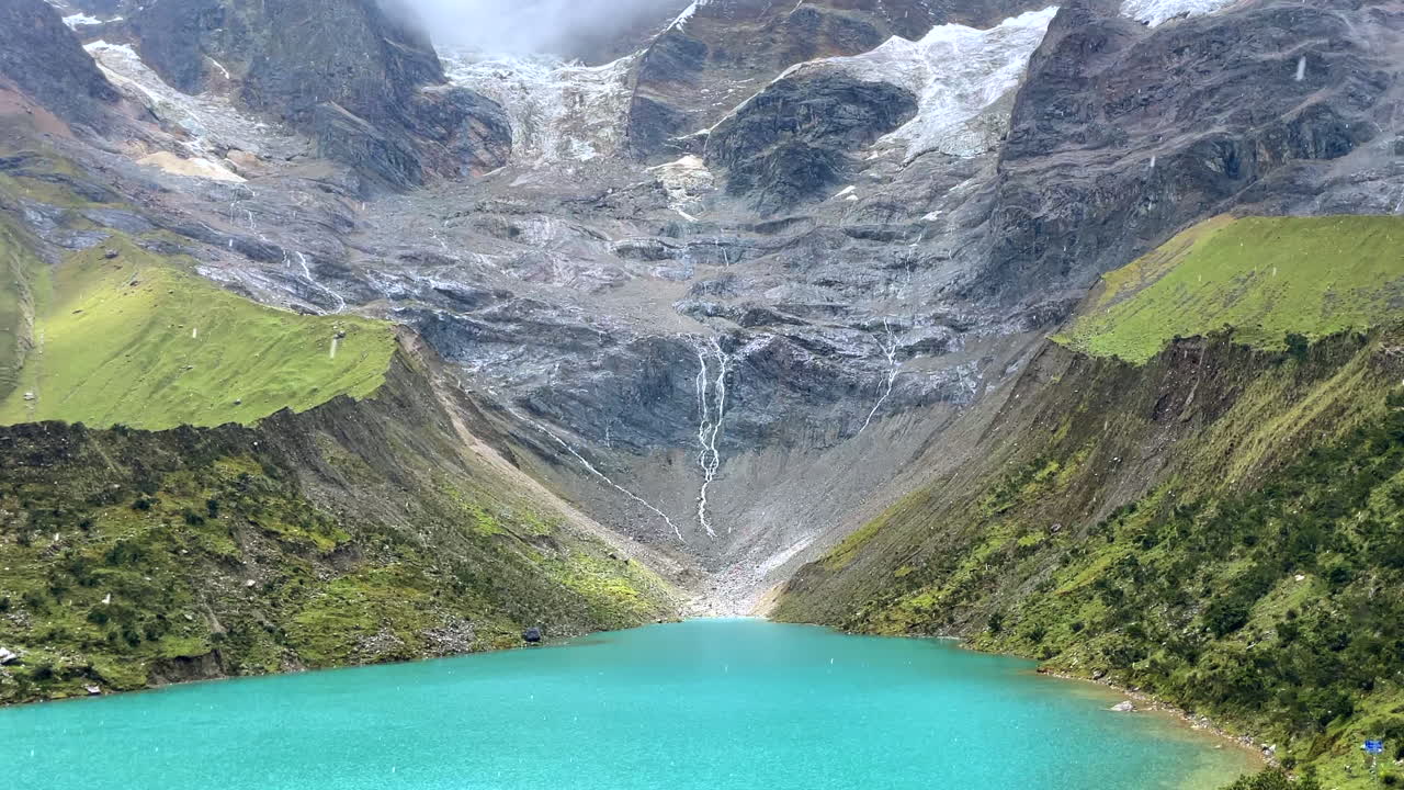 Zoom indietro da una laguna turchese sotto la pioggia con lo sfondo innevato delle Ande Salkantay