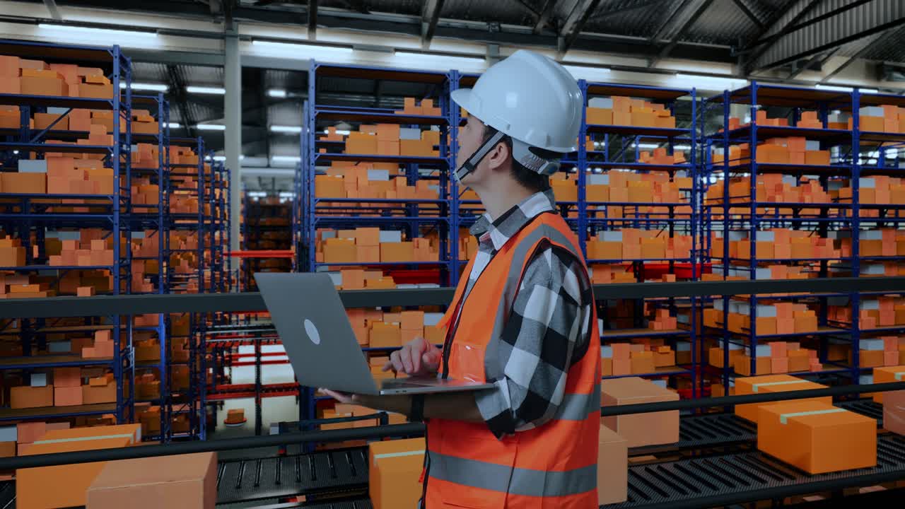 vista lateral de un ingeniero masculino asiático con casco de seguridad mirando una computadora portátil y mirando a su alrededor mientras está de pie en el almacén con estantes llenos de productos de entrega