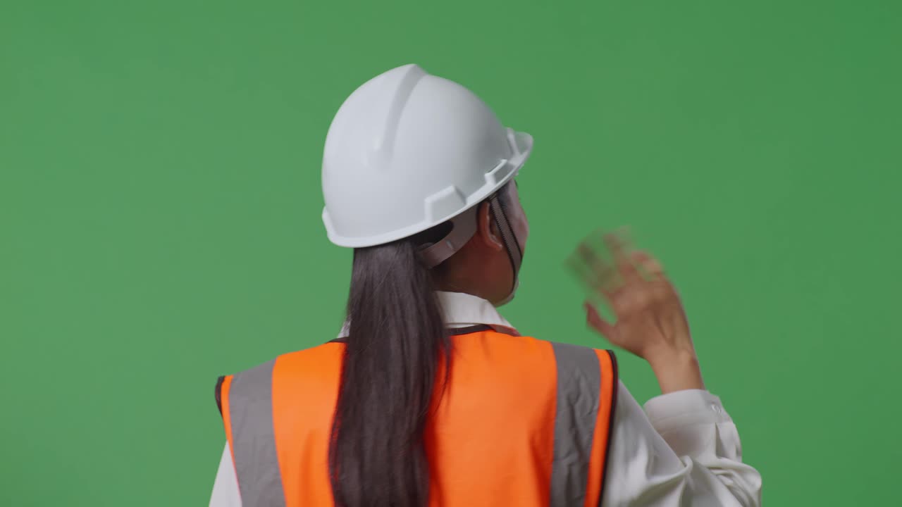 안전 헬을 입은 아시아 여성 엔지니어가 녹색 스크린 배경 스튜디오에서 걸어가는 동안 손을 흔들고 있는 근접한 뒷면