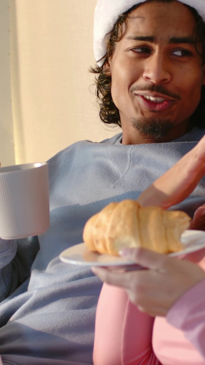 Vertical video: Reacting to croissant offering on sofa, Santa hat man refusing pastry holding mug
