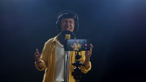 young asian boy with headphone singing into a condenser microphone while recording video by smartphone on the black background