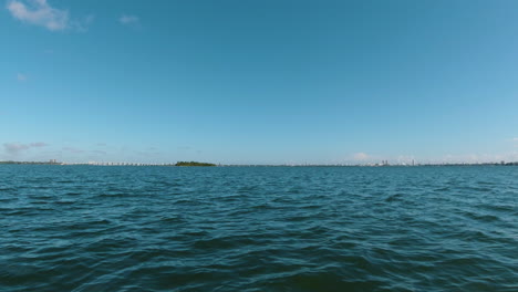 Punto-De-Vista-De-Un-Pequeño-Bote-De-Cielo-Azul-Se-Encuentra-Con-Agua-Azul-Con-Una-Pequeña-Isla-En-El-Horizonte