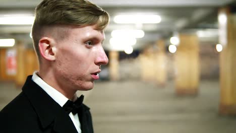 Close-Up-view-of-young-blonde-man-in-a-black-suit-with-a-bow-tie-smoking-a-cigarette-in-the-parking.-Waiting-for-someone