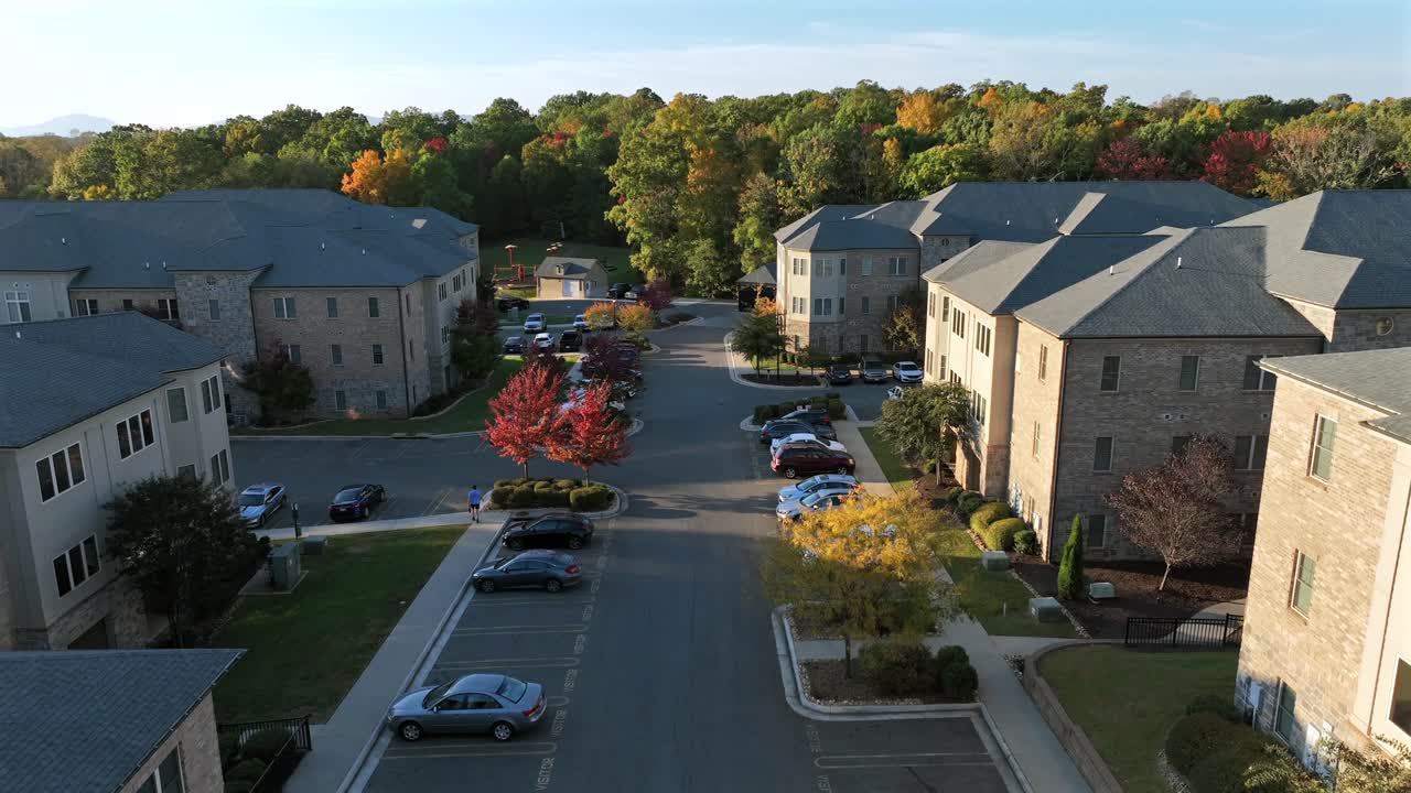 Luxury apartment houses in suburb of american town with parking cars. Beautiful sunny day autumn day with colorful trees in suburbia. Rising drone wide shot. Silhouette of mountains in background.