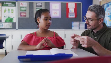 un maestro de sexo masculino diverso mostrando ejercicios a una estudiante sentada en un escritorio en el aula