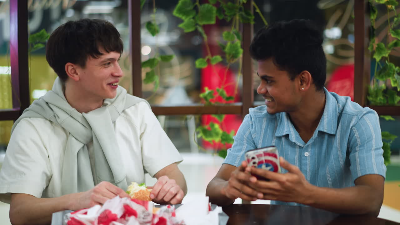Light skinned young man pointing at phone while laughing with friend during casual interaction, sharing funny content, smiling faces and lively expression, closeup social moment with digital device