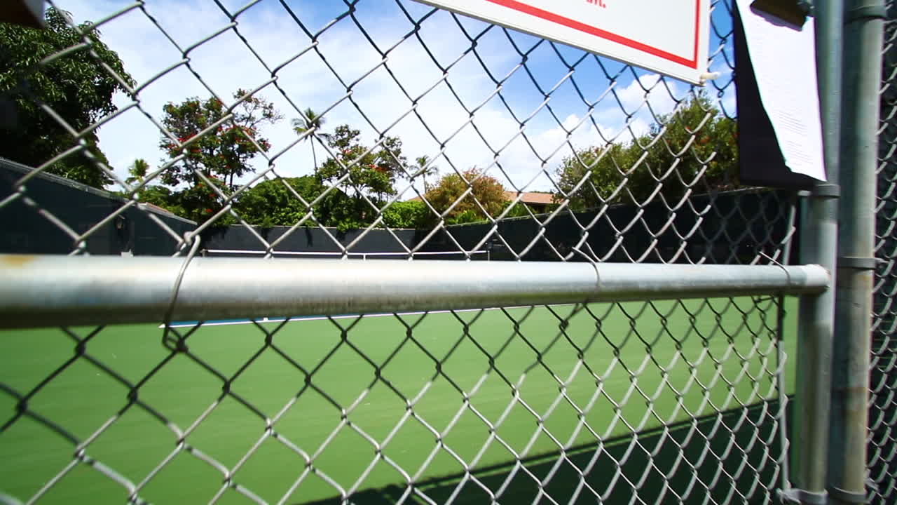 hand-held shot of Tennis court viewed through a chain-link fence.