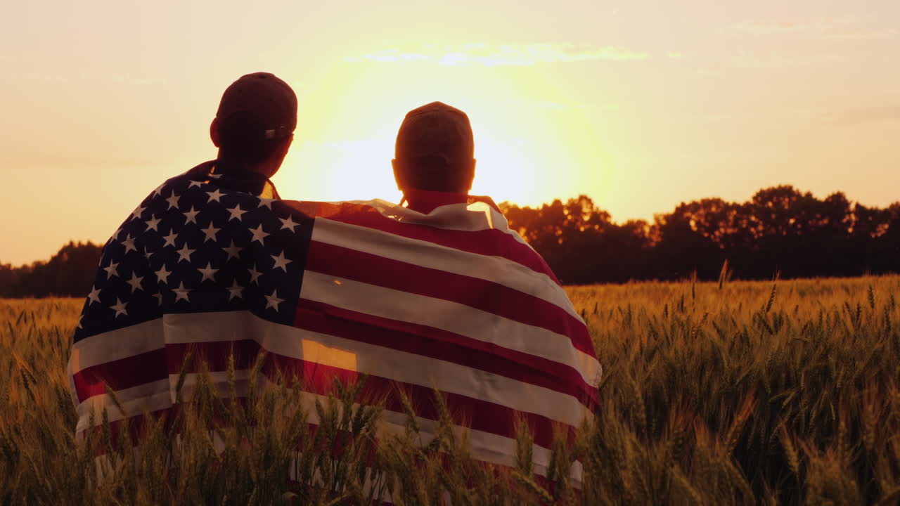 dos hombres con la bandera de américa sobre sus hombros miran el amanecer sobre un campo de trigo