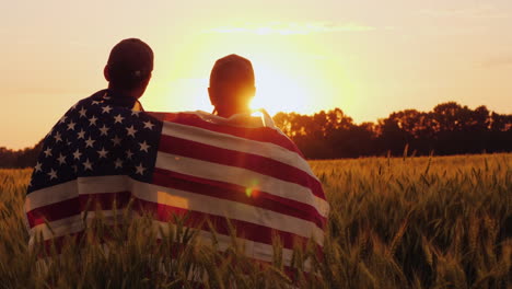 two men with the flag of america on their shoulders look at the sunrise above a wheat field