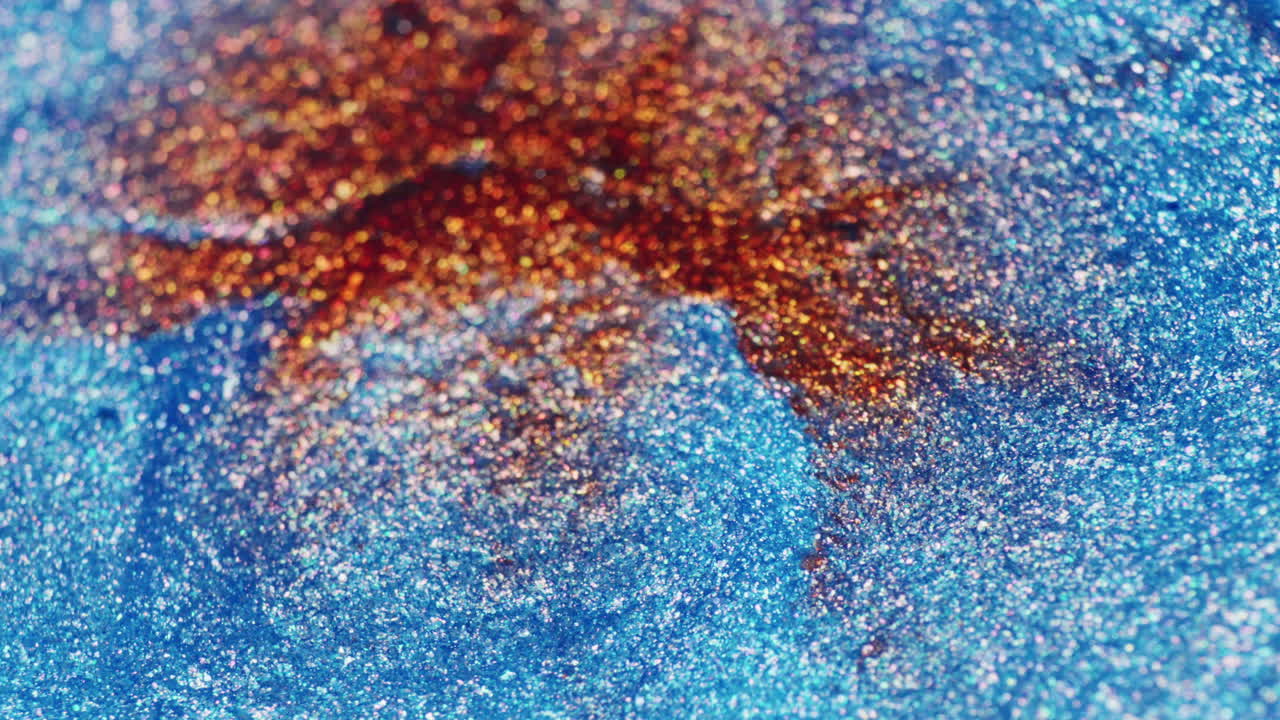 A water drop hits glittery red and blue ink in macro slow motion, creating ripples and shimmering movement