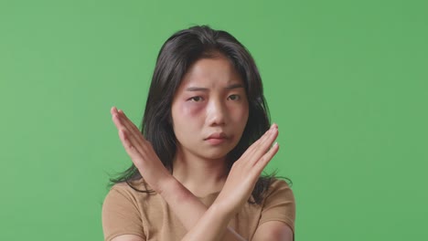 close up of young asian woman with bruise on face looks into camera showing hands cross to stop violence in the green screen background studio