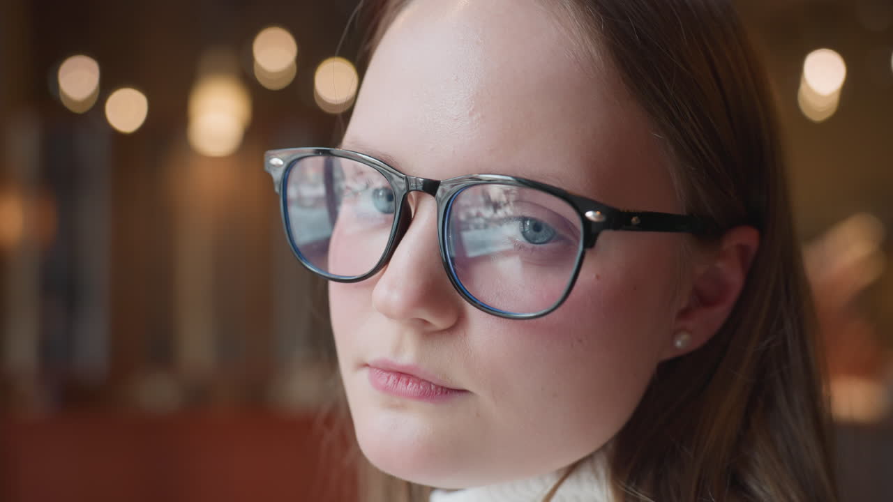 young woman in glasses gazes out window then turns toward camera with calm expression, surrounded by soft indoor lighting and cozy ambiance, reflecting thoughtful mood in serene environment