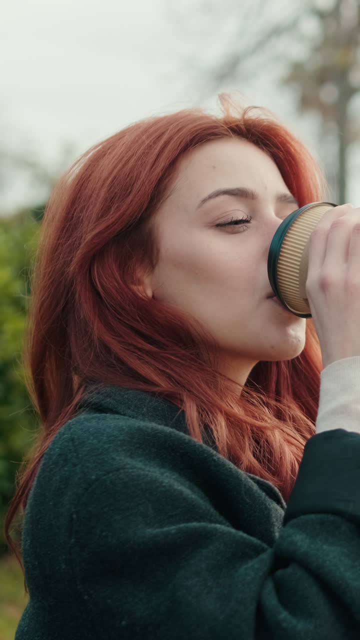 Ung rødhåret kvinde drikker kaffe udendørs i en park