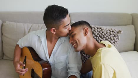 pareja de hombres homosexuales de raza mixta sonrientes sentados en el sofá y besándose mientras uno toca la guitarra