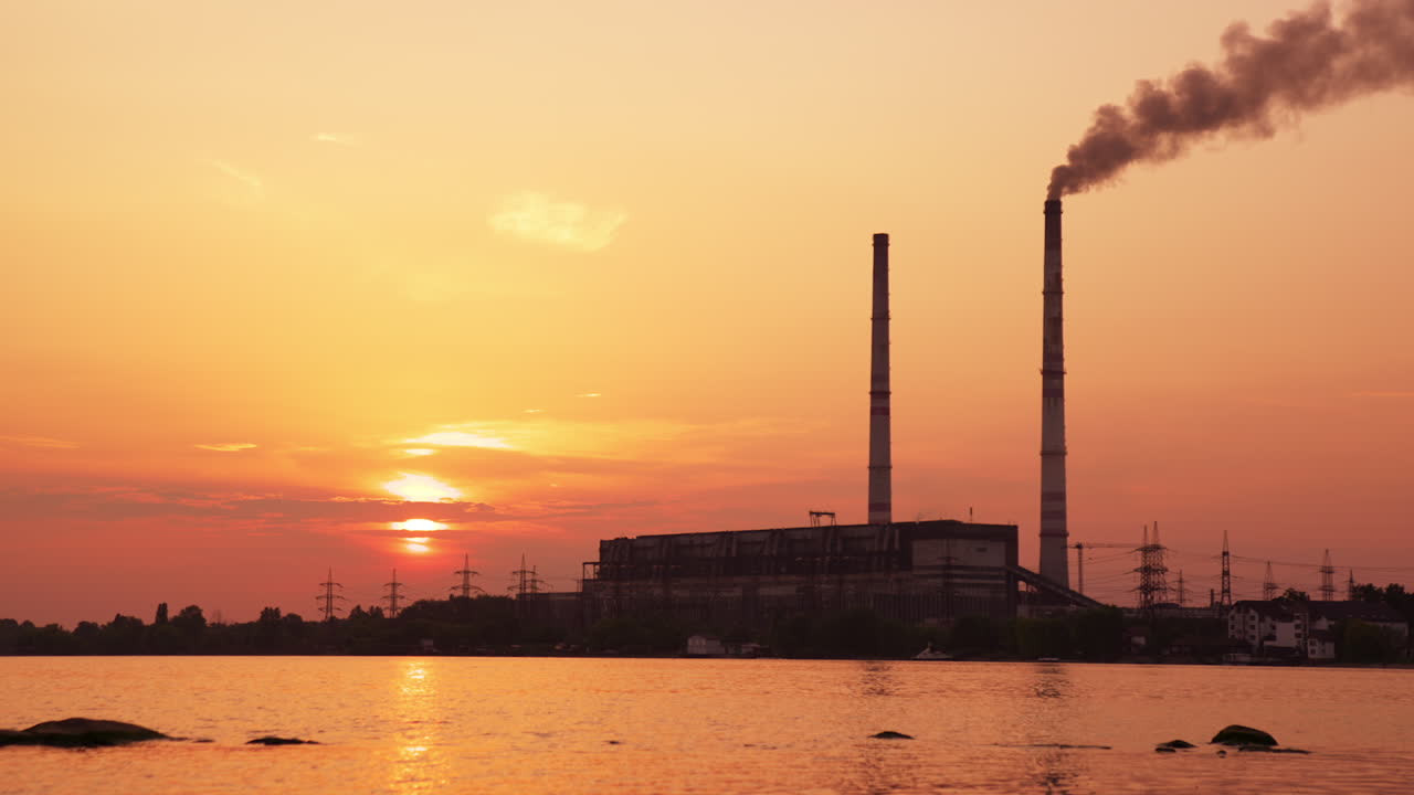 Bright setting sun hiding in the clouds at sunset. Industrial plant working near the river producing thick smoke from high pipe.