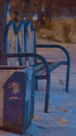banco vacío rodeado de nieve bajo una iluminación suave con figuras borrosas caminando en el fondo lejano, creando un entorno pacífico de la noche de invierno con colores y sombras serenos y apagados