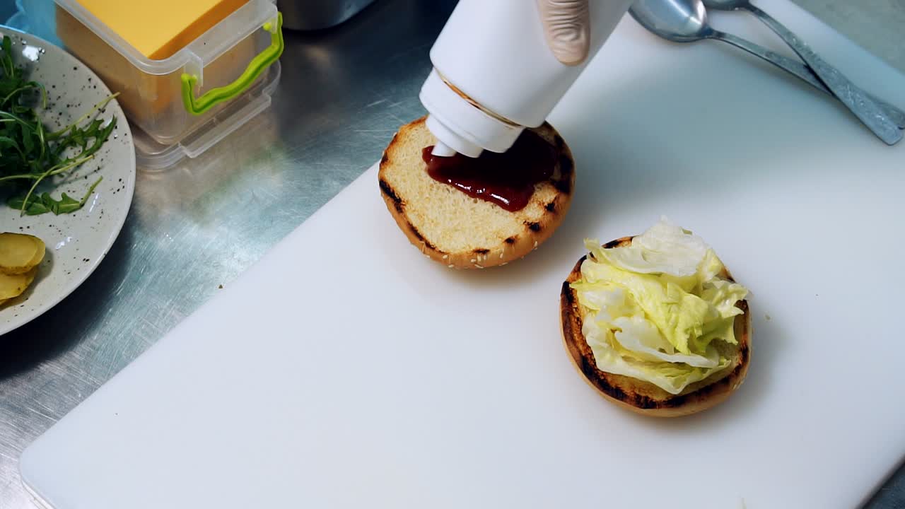 Burger cooking. Cook pours red sauce on a burger bun. Chef preparing juicy burger and pours spicy tomato sauce on it making burgers at fast food restaurant