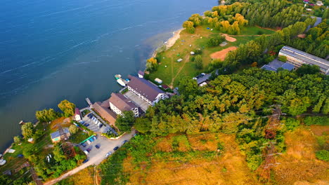 Hotel located on the river bank with few piers and boats. Green waterfront and dam at backdrop. Aerial view.