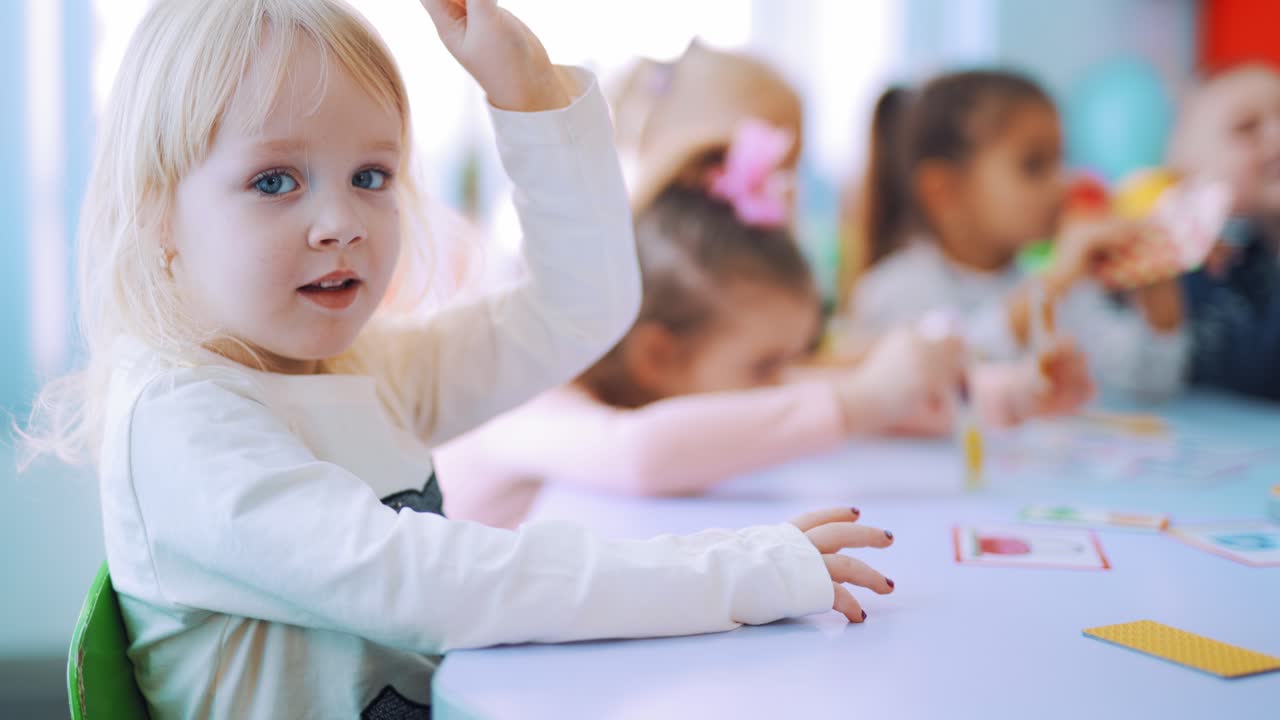 Small girl with a card on the background of schoolchildren during the lesson. Cute child with blond hair is sitting at the table and raising one picture up while looking at camera.
