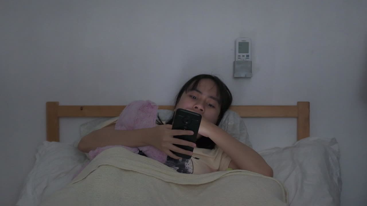 Southeast asian woman sits in bed yawns while scrolling on phone, dozes off to sleep