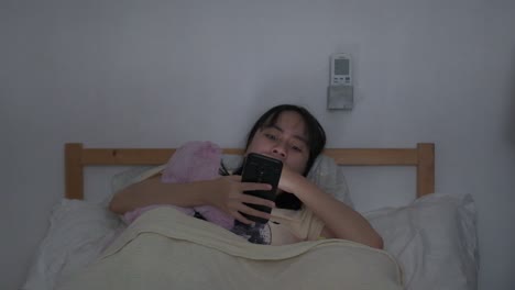 southeast asian woman sits in bed yawns while scrolling on phone, dozes off to sleep