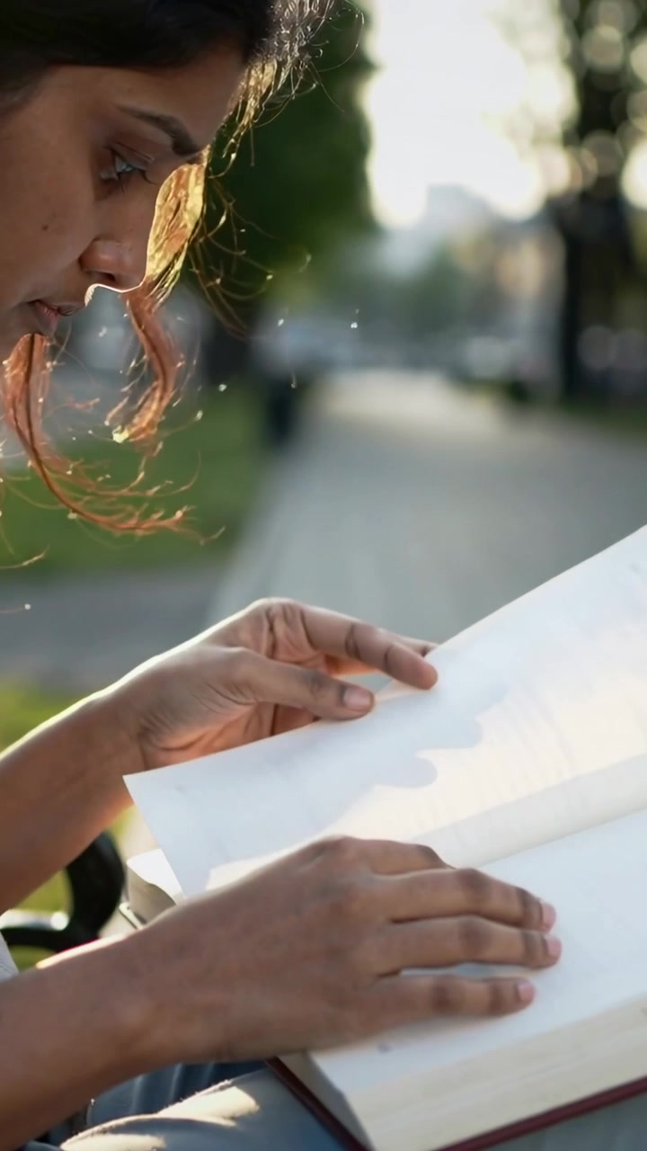 Vertical video: Looking down, woman in T-shirt turning pages and reading book on bench, copy space