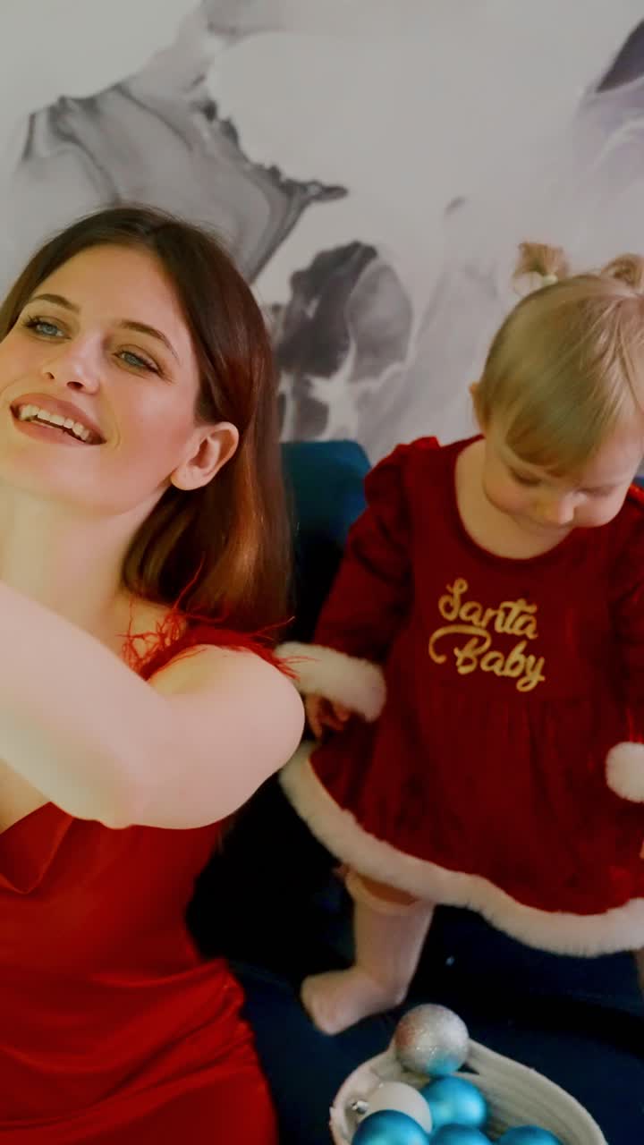 A Joyous Holiday Moment: A Mother Interacts with Her Young Daughter, Dressed in a Festive Red Dress, Sharing Laughter and Decorating with Colorful Ornaments on a Cozy Sofa