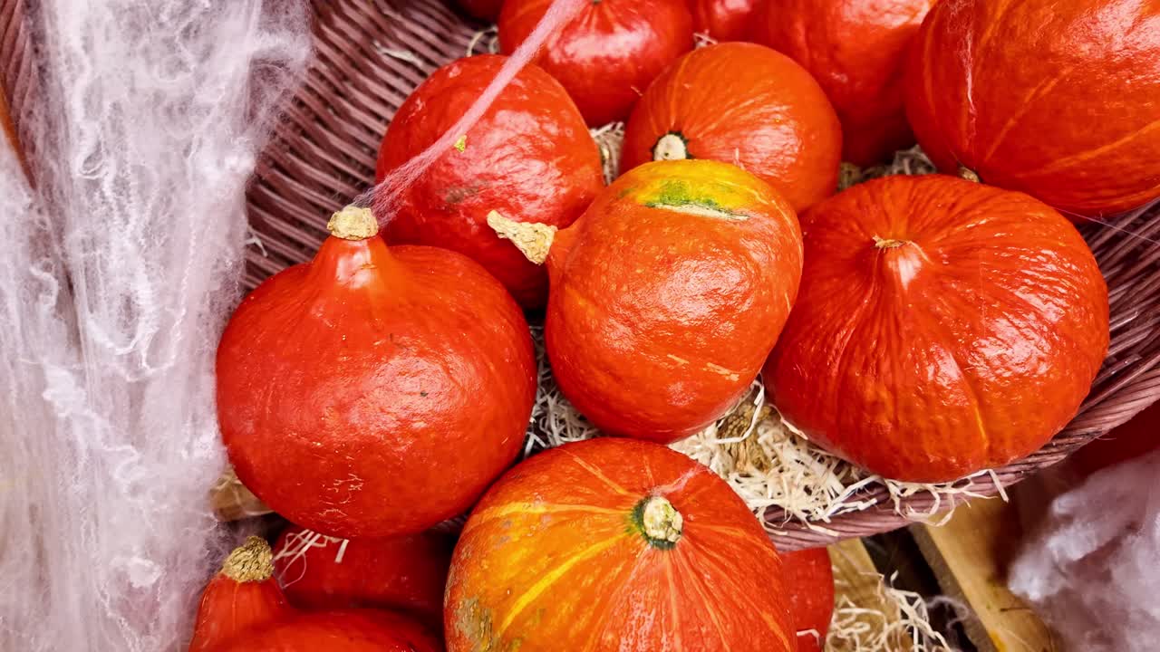 Top-down close-up shot moving over several red kuri squash arranged in baskets with straw and decorative spider webs on a market display