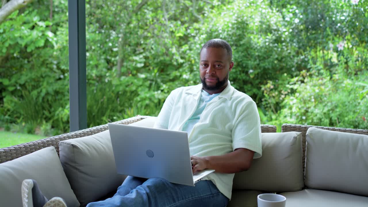 Adult African man reclining at veranda, typing reply on silver laptop, laughing at funny message