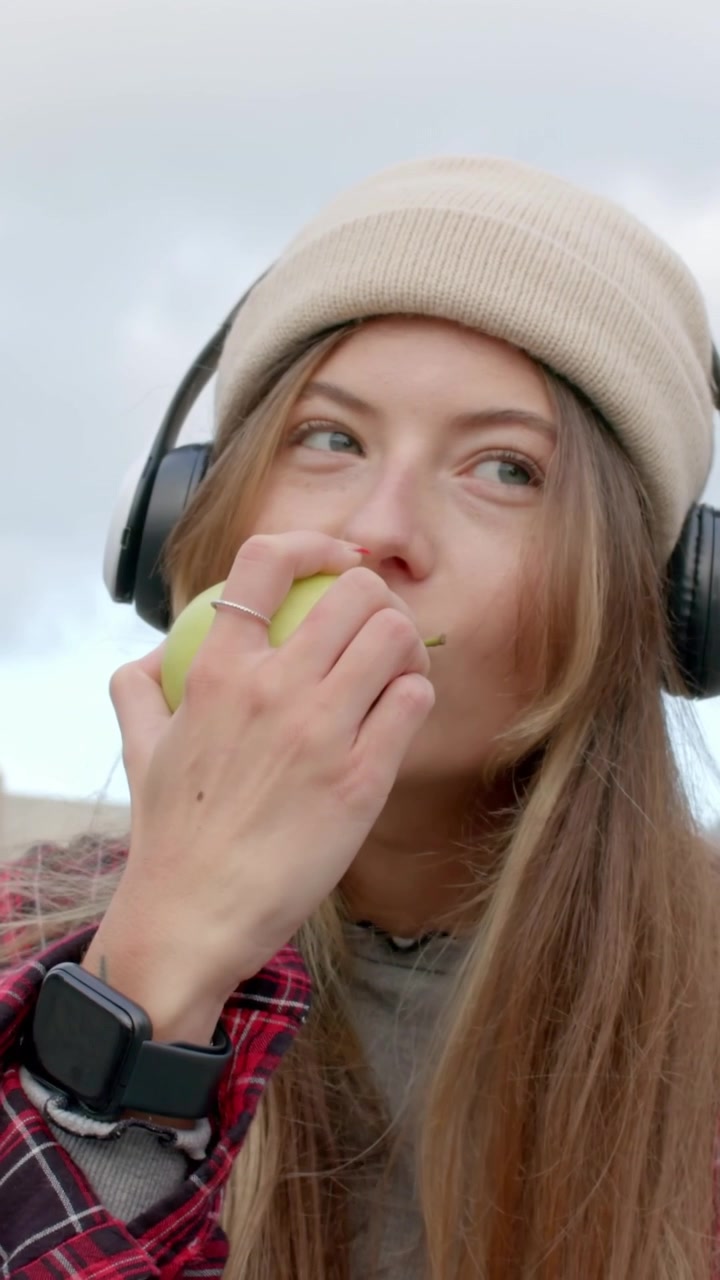 Video verticale: Donna con berretto e cuffie che mangia una mela verde sul tetto.
